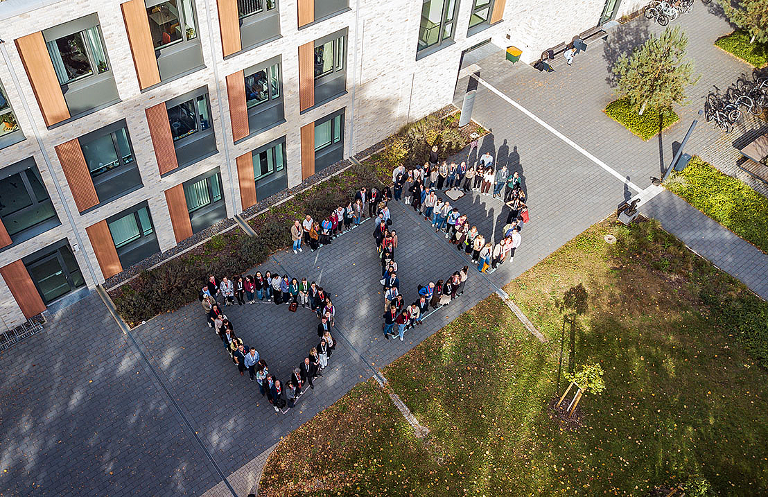 Luftaufnahme von einer Gruppe Menschen, die auf einem gepflasterten Platz vor einem Gebäude die Buchstaben "DZD" bilden