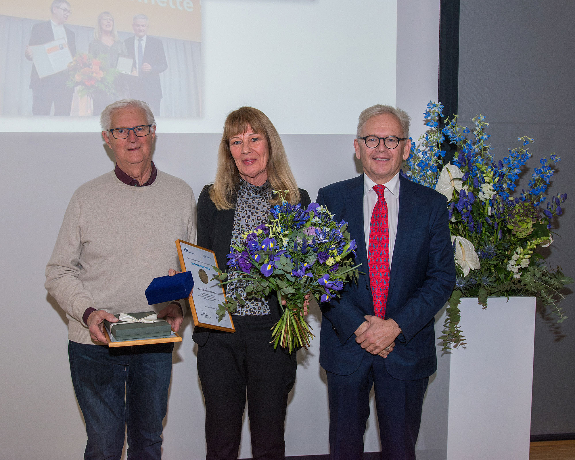 Drei Personen stehen nebeneinander, die mittlere Frau hält einen Blumenstrauß und eine Urkunde, der Mann links hält eine Schachtel und ein Buch, der Mann rechts trägt einen Anzug mit roter Krawatte.