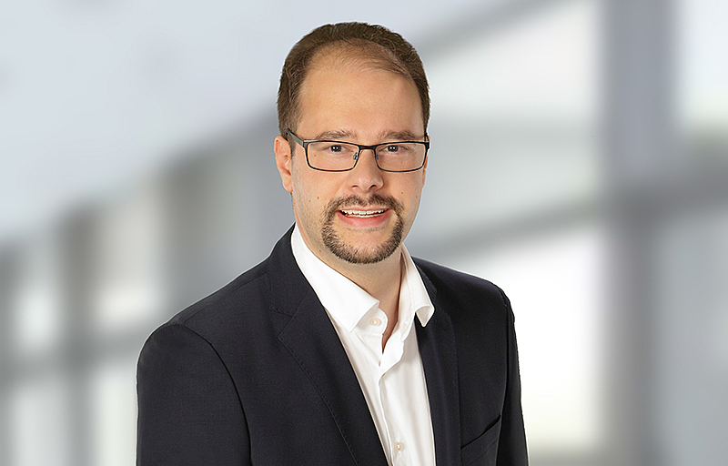 Porträt eines Mannes mit Brille, kurzem Haar und Bart, der einen dunklen Anzug und ein weißes Hemd trägt, vor einem unscharfen hellen Hintergrund